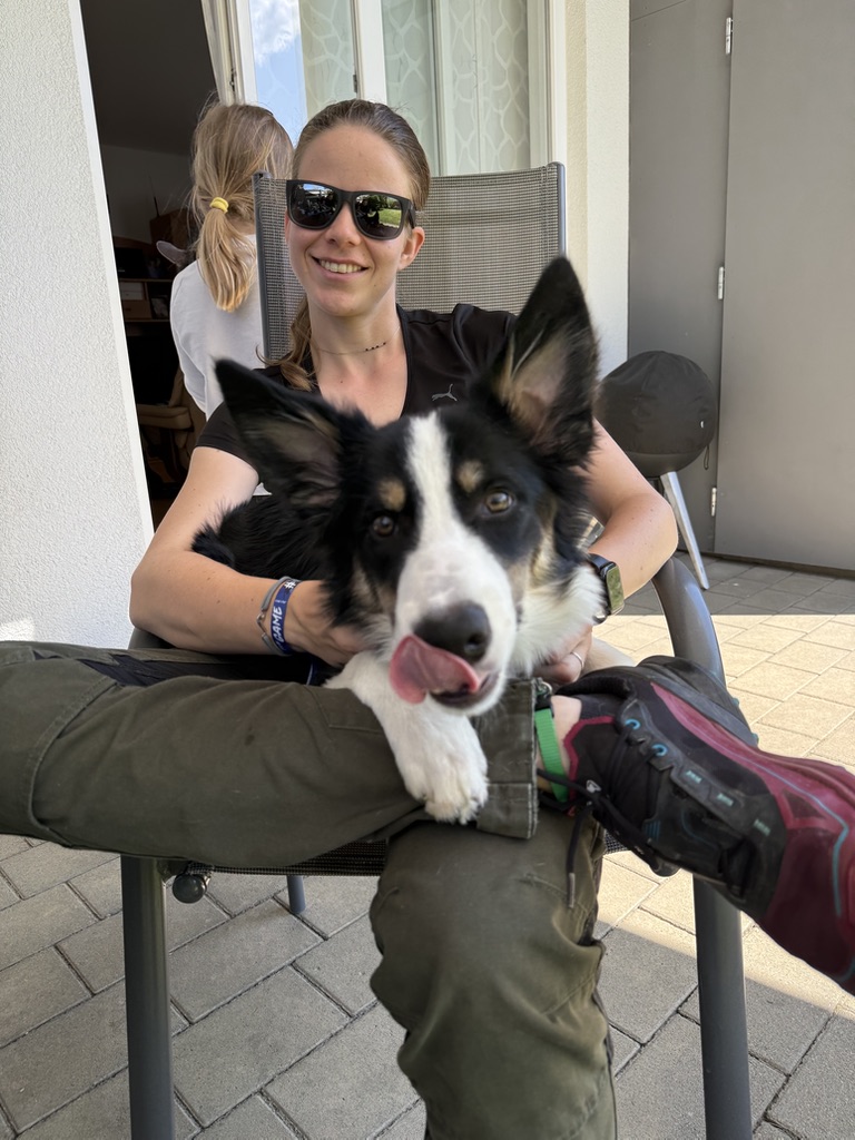 Eine Frau sitzt am Sessel und hat einen Hund am Schoß der sich über die Schnauze schleckt.