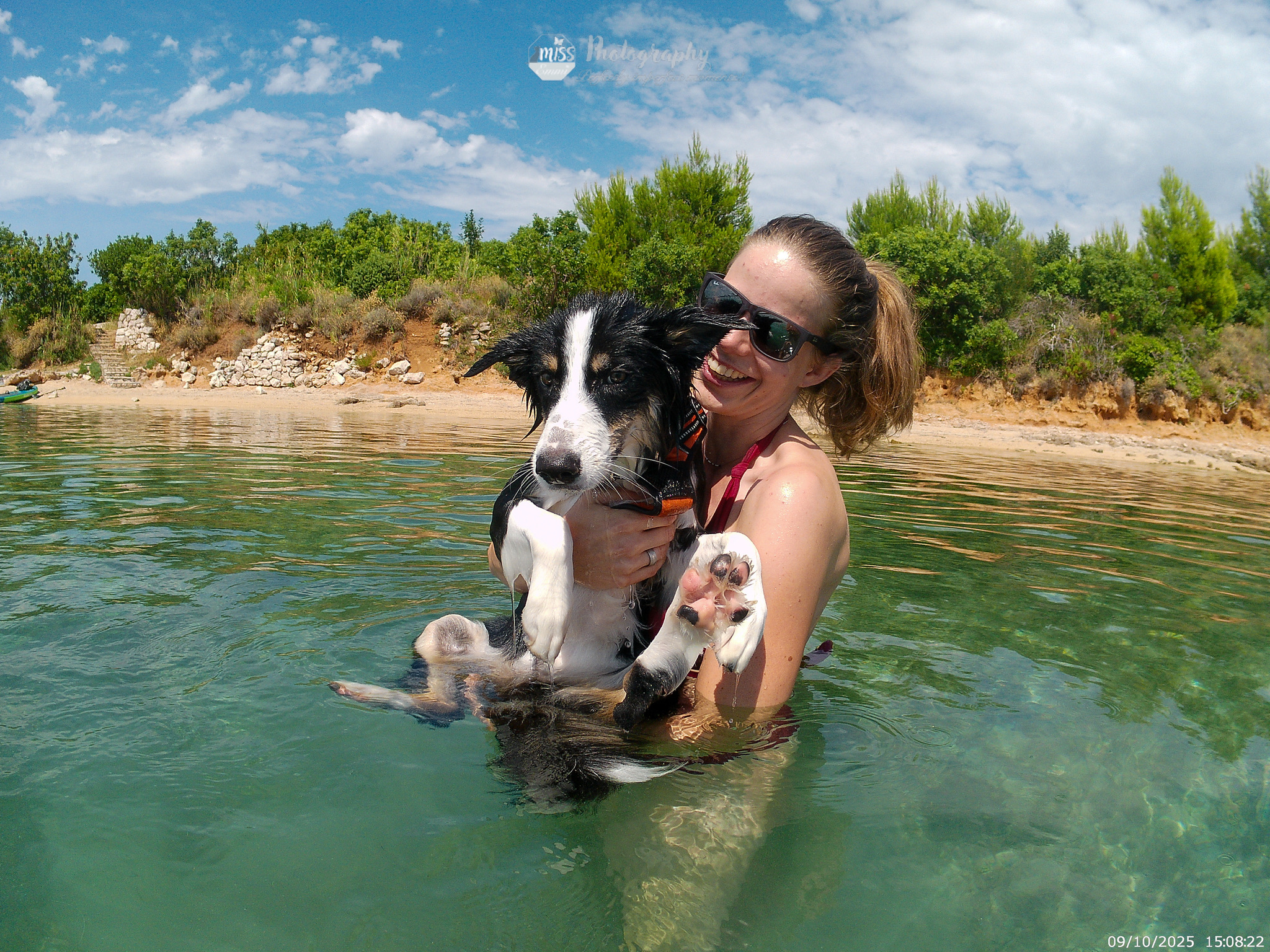 Eine Frau mit Sonnenbrille steht im Wasser und hat einen Welpen im Arm der ganz nass ist.