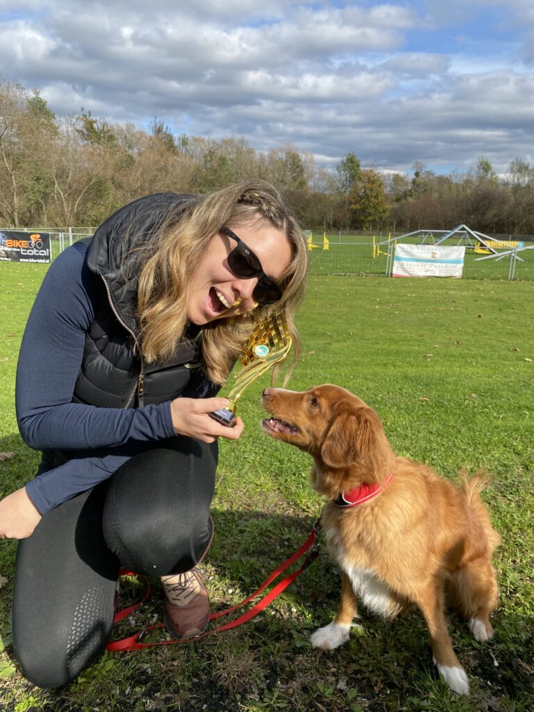 Eine Frau mit einem Pokal in der Hand kniet in der Wiese und neben ihr eine Nova Scotia Duck Tolling Retriever Hündin
