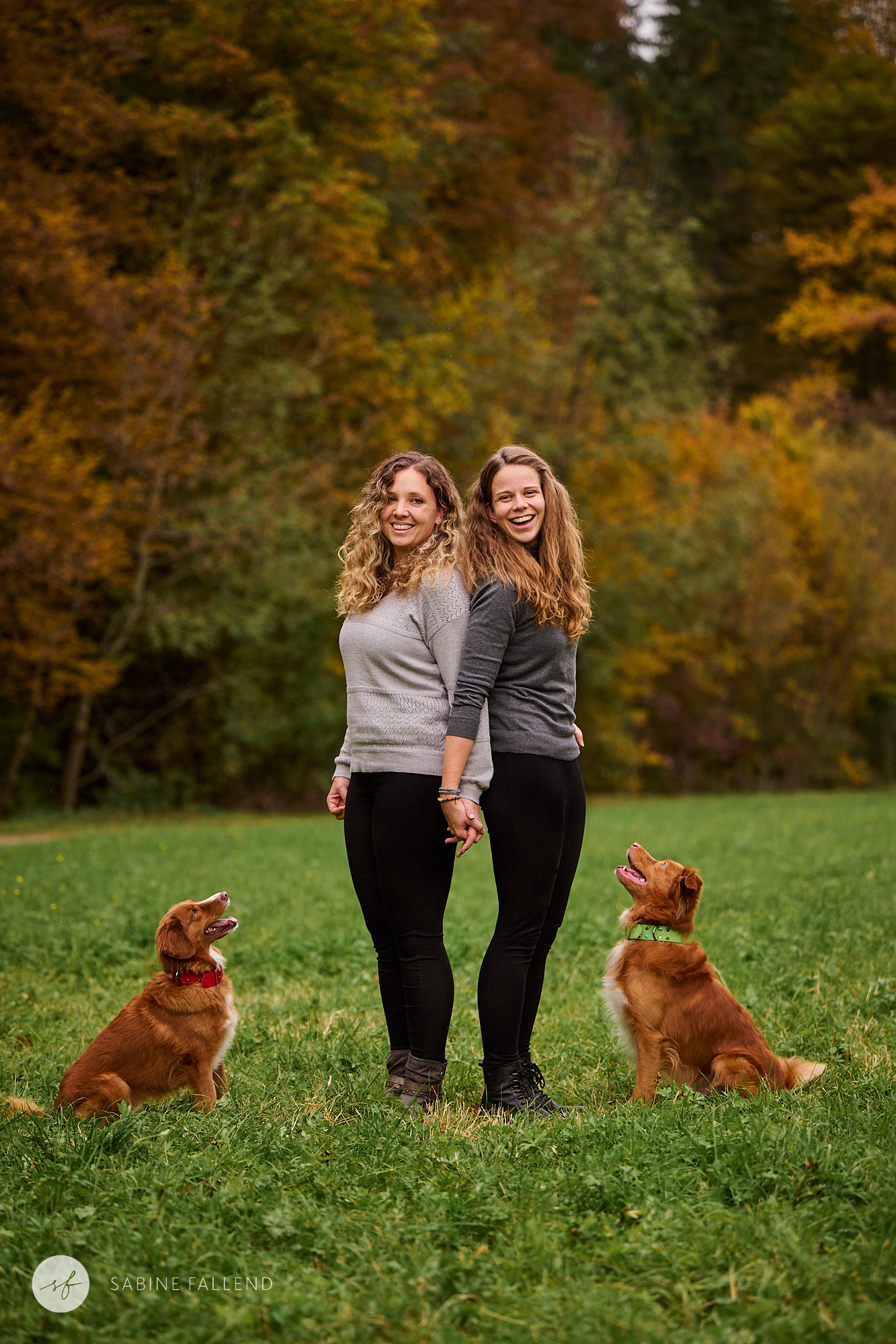 Zwei Frauen stehen mit dem Rücken zueinander und ihre Hunde sitzen jeweils vor ihnen