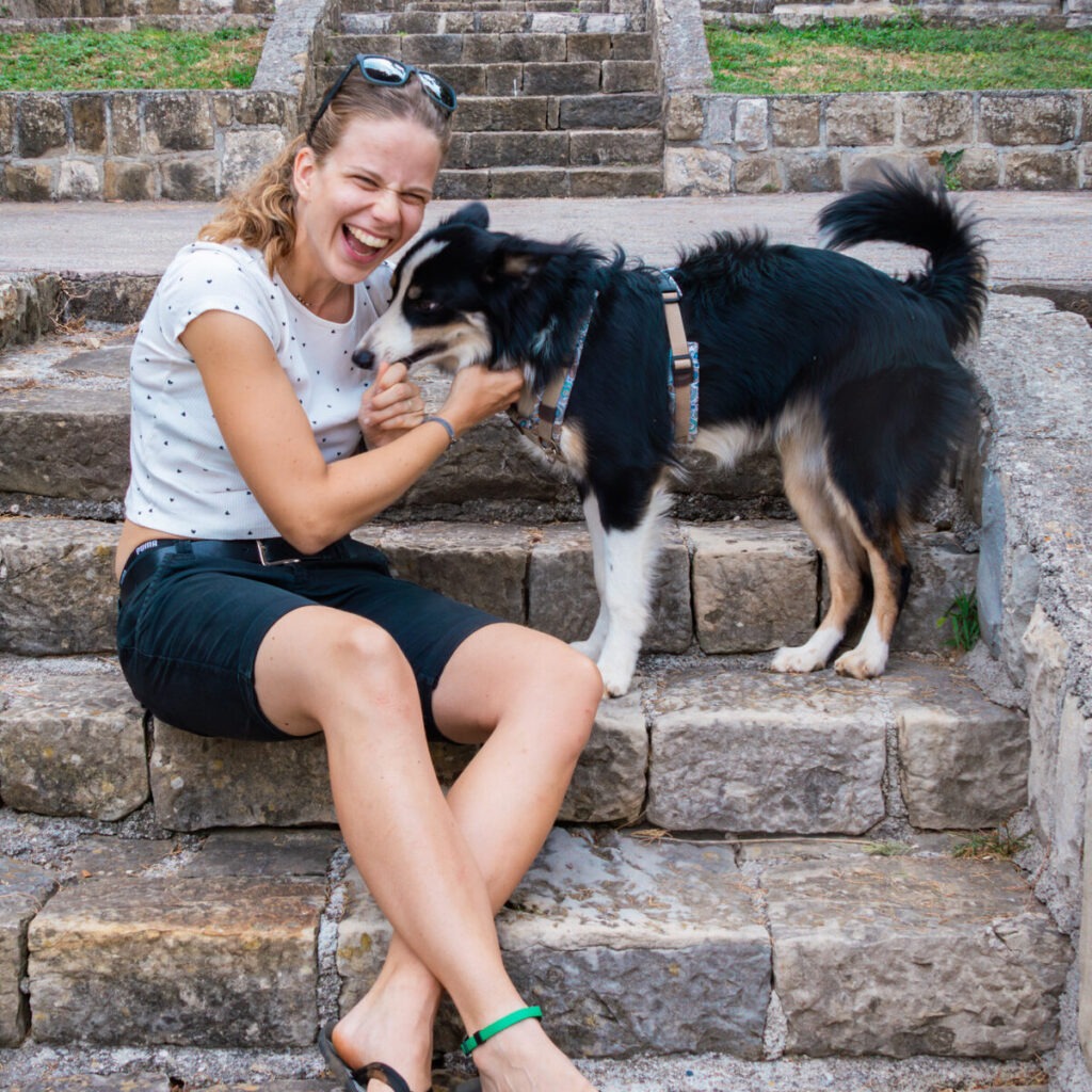 Eine Frau sitzt auf einer Steinstiege und lacht herzlich während sie einen Hund streichelt.
