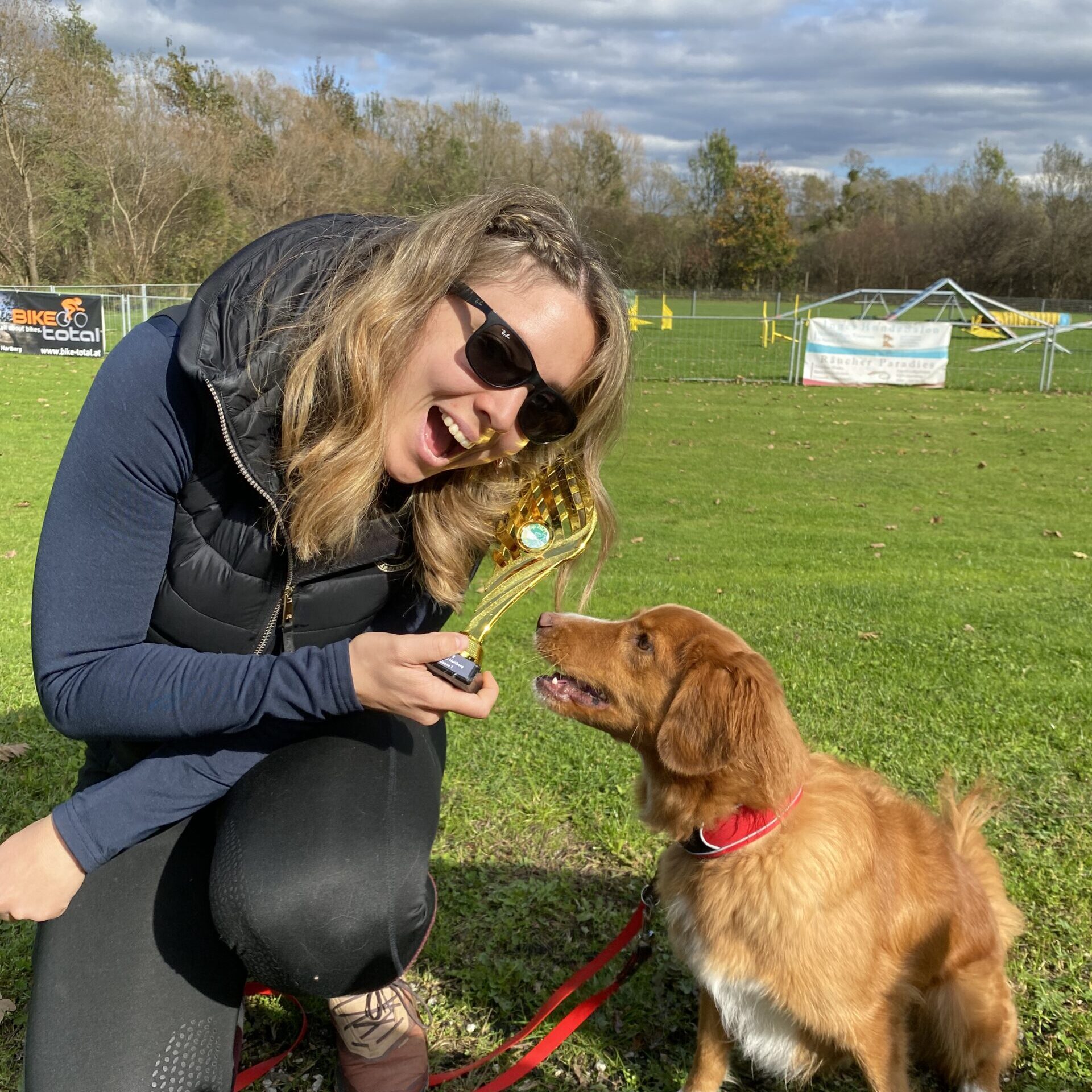 Eine Frau mit einem Pokal in der Hand kniet in der Wiese und neben ihr eine Nova Scotia Duck Tolling Retriever Hündin